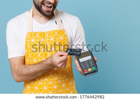 Cropped image of young male chef or cook baker man in apron white t-shirt isolated on blue background. Cooking food concept. Hold bank payment terminal to process and acquire credit card payments