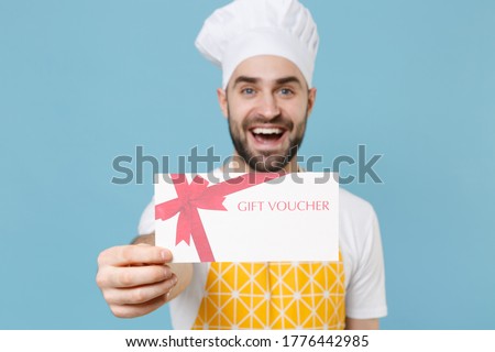 Cheerful young bearded male chef or cook baker man in apron white t-shirt toque chefs hat isolated on blue background studio portrait. Cooking food concept. Mock up copy space. Hold gift certificate
