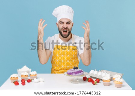 Angry young bearded male chef or cook baker man in apron white t-shirt toque chefs hat cooking at table isolated on blue background in studio. Cooking food concept. Spreading hands screaming swearing