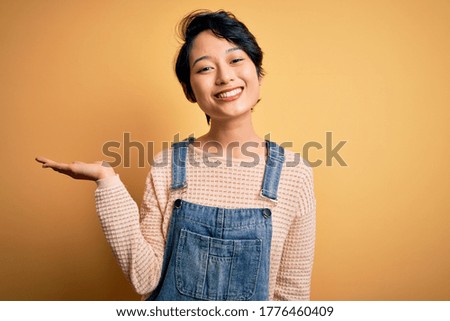 Young beautiful asian girl wearing casual denim overalls over isolated yellow background smiling cheerful presenting and pointing with palm of hand looking at the camera.