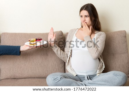 Pregnant woman sitting on the sofa don't want to eat a cake and makes stop gesture. No sweet and dessert during pregnancy concept.