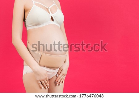 Close up of supporting orthopedic bandage against backache on pregnant woman in underwear at pink background with copy space. Orthopedic abdominal support belt concept.
