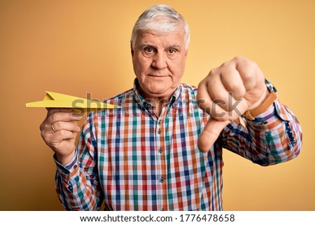 Senior handsome hoary man holding paper airplane standing over isolated yellow background with angry face, negative sign showing dislike with thumbs down, rejection concept
