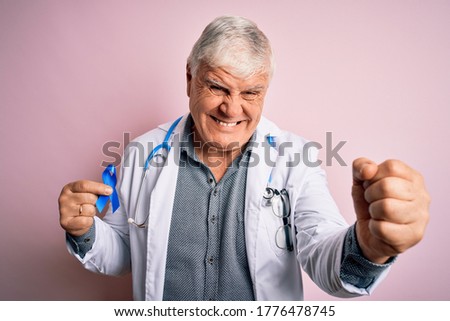 Senior handsome hoary doctor man wearing stethoscope holding blue cancer ribbon annoyed and frustrated shouting with anger, crazy and yelling with raised hand, anger concept