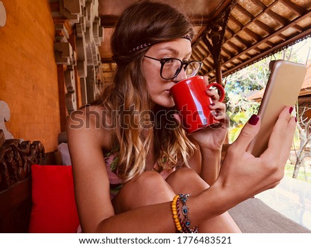 Modern young woman using cellphone and drinking coffee on a terrace sofa.