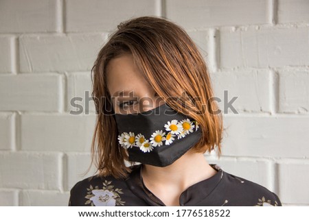 young woman in the black medical mask with chamomile against the background of white bricks. Creative medical mask with flowers for corona virus protection.