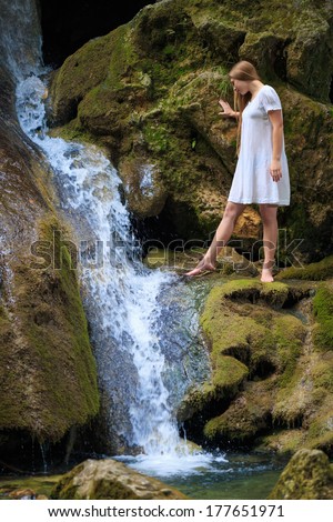 Beautiful young woman with flowing hair in white sundress stands above the waterfall on mossy rock