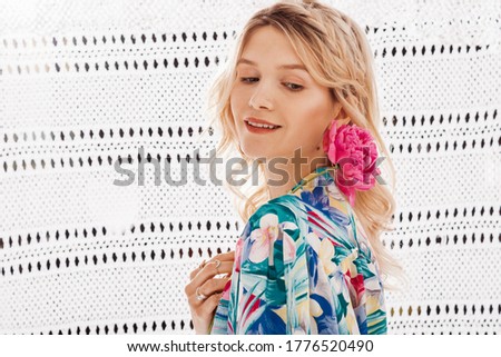 Close up portrait of young beautiful blonde happy smiling wearing trendy blue earrings, many rings, summer dressing gown, holding pink peony flower. Copy, empty space for text