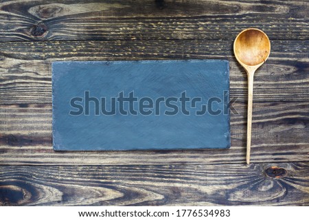 Old handcrafted wooden spoon with deep bowl made of olive wood lying near a slate stone over a rustic wood table background. Image shot from top view. Flatlay.