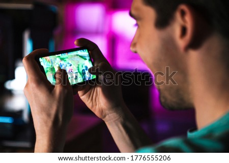 Image closeup of smiling man playing video game on mobile phone and computer at home