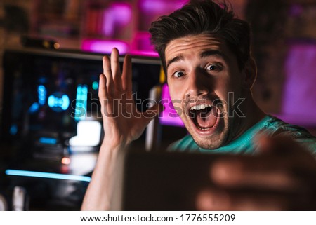Image of delighted man screaming and taking selfie photo on cellphone while playing video game at home