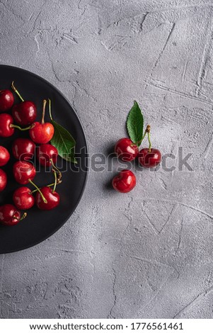 Fresh ripe cherry fruits with green leaves on black plate, summer vitamin berries on grey stone background, top view