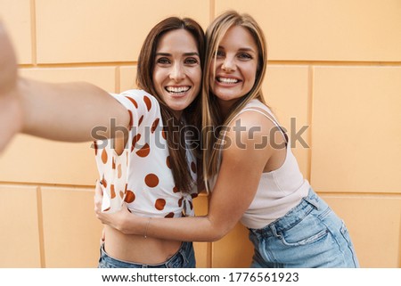 Image of cheerful adult two women taking selfie photo and hugging while posing on city street