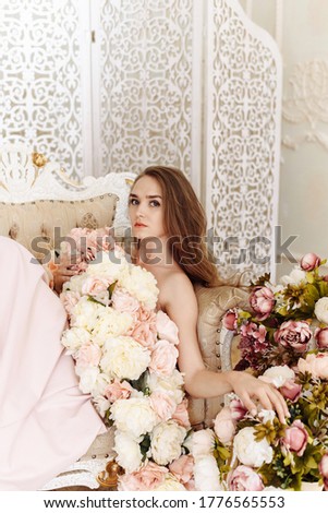 Beauty, fashion. Wedding style. Portrait of a beautiful bride girl in an elegant white dress of roses, peonies and flowers, sitting on a sofa in the interior