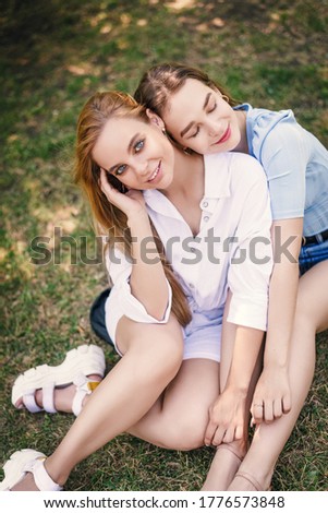 Two beautiful girls have fun in the park on a sunny summer day. The sisters are sitting on a green lawn, laughing.