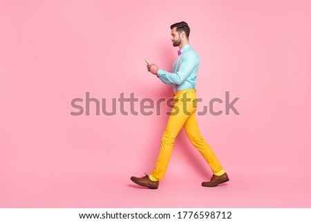 Full length profile photo of handsome guy trend clothes browsing telephone writing new post walk street wear shirt suspenders bow tie yellow trousers isolated pastel pink color background