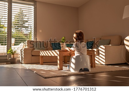 Light and shadows. Little toddler boy sit on potty playing video game on digital tablet in living room at home. Beautiful interior