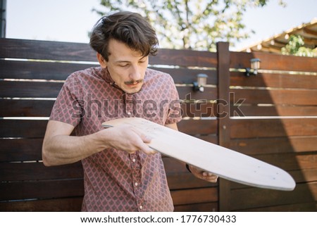 Man blowing wood dust from polished skateboard deck outdoor. Brunette man cleaning wooden board from dust while blowing.
