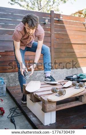 Young man using polish machine in outdoor workshop. Male carpenter holding power sander while standing outdoor next to pallet with skateboard.
