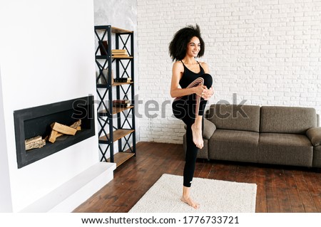 Warm up before training at home. An African-American beautiful girl in sportswear is doing stretching legs in cozy living room