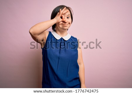 Young down syndrome woman wearing elegant shirt over pink background doing ok gesture with hand smiling, eye looking through fingers with happy face.