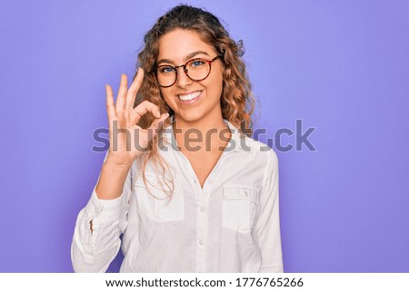 Young beautiful woman with blue eyes wearing casual shirt and glasses over purple background smiling positive doing ok sign with hand and fingers. Successful expression.