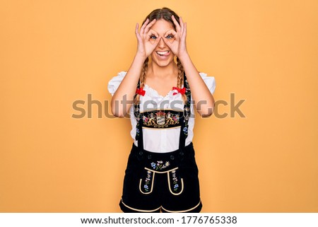 Young beautiful blonde german woman with blue eyes wearing traditional octoberfest dress doing ok gesture like binoculars sticking tongue out, eyes looking through fingers. Crazy expression.