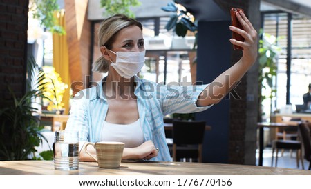 Happy young woman taking selfie in the cafe. Portrait of a happy woman taking a photo with modern smartphone. Casual cheerful woman taking selfie smiling. Woman wearing a protective mask.