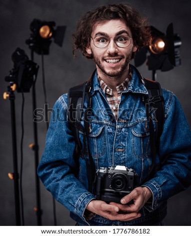 Young curly hipster man with curly hairstyle in glasses wearing denim jacket holds a camera. Studio portrait with lighting equipment in the background