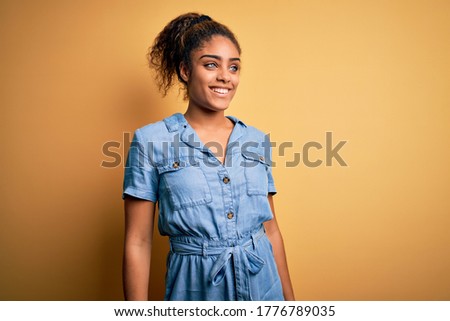 Young beautiful african american girl wearing denim dress standing over yellow background looking away to side with smile on face, natural expression. Laughing confident.