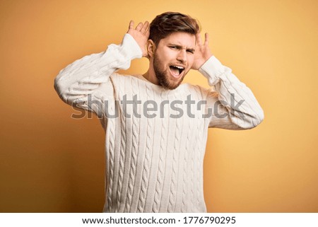 Young blond man with beard and blue eyes wearing white sweater over yellow background Smiling cheerful playing peek a boo with hands showing face. Surprised and exited