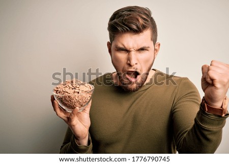 Young blond man with beard and blue eyes holding bowl with cornflakes cereals annoyed and frustrated shouting with anger, crazy and yelling with raised hand, anger concept