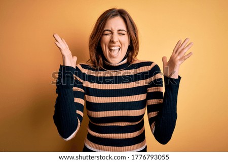 Young beautiful brunette woman wearing striped turtleneck sweater over yellow background celebrating mad and crazy for success with arms raised and closed eyes screaming excited. Winner concept