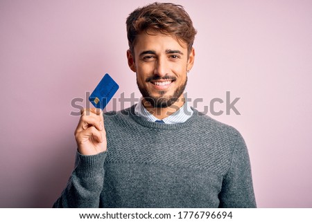 Young customer man with beard holding credit card for payment over pink background with a happy face standing and smiling with a confident smile showing teeth