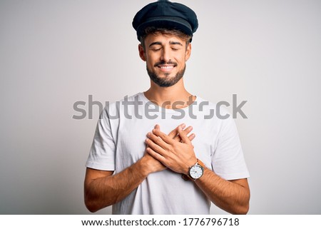 Young driver man with beard wearing hat standing over isolated white background smiling with hands on chest with closed eyes and grateful gesture on face. Health concept.