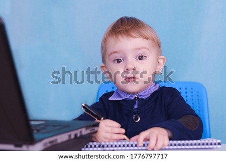 little blond boy sits at a laptop with a pen and makes notes. Emotions of surprise and joy on the face. Blue background. business concept.
