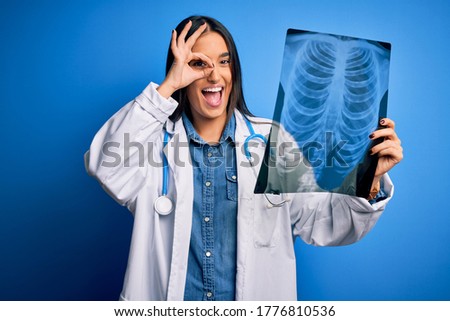 Young beautiful brunette doctor woman wearing stethoscope holding chest xray with happy face smiling doing ok sign with hand on eye looking through fingers