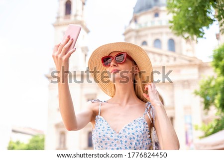 Portrait shot of happy young woman wearing straw hat and using mobile phone while making selfie. Background with the St. Stephen’s Basilica in Budapest.