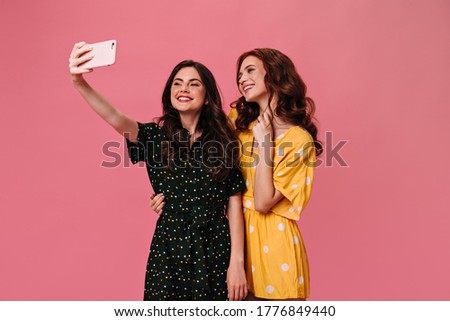 Charming women in polka dot outfits taking selfie on pink background. Two beautiful girlfriends in beautiful dresses hugging on isolated