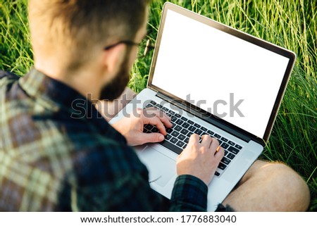 Young hipster student using laptop on grass in park at sunset. Blank white screen copy space. View over the shoulder.
