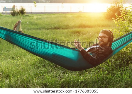 Cheerful handsome bearded male hipster in round eyeglasses  and headphones lying i hammock smiling and looking to camera in park at sunset. Sun flare.