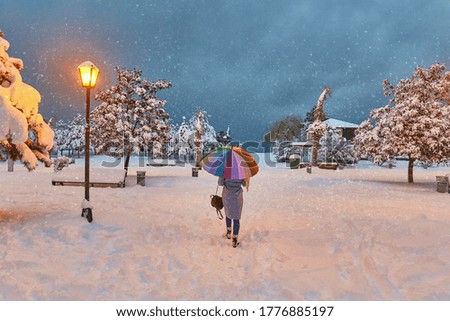 Alone lonely woman with colorful umbrella walks in the quiet winter park in the evening time. Winter bewitching fairytale park with flying snowflakes