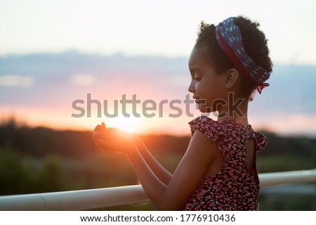 Portrait of a girl at sunset. Child holds the sun in her hand. Horizon, landscape on the background. Mixed race person, afro hair. Concept of happiness, dreams, hope, vacation, life, nature. Royalty-Free Stock Photo #1776910436