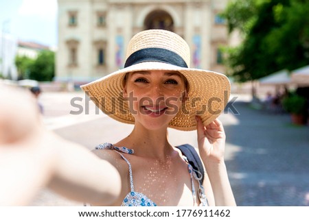 Close-up shot of cute young girl is making selfie on a camera while walking on the street. She is wearing straw hat. 