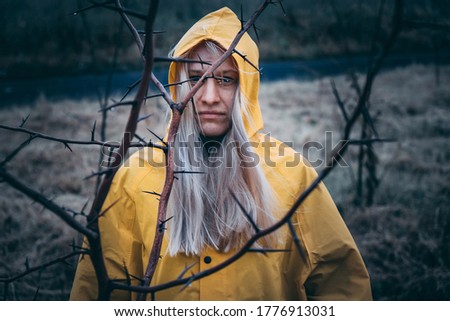 a girl in a yellow cloak in cloudy rainy weather looks with a worried look through the branches of a tree with thorns. hopelessness concept