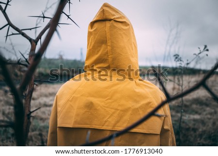 back of a man in a yellow cloak in cloudy rainy weather through the branches of a tree with thorns. trouble expectation concept
