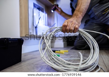 Close up shot of hand of aged electrician repairman in uniform working, fixing, installing ethernet cable in fuse box, holding flashlight and cable. Selective focus on hand and cable. Horizontal shot Royalty-Free Stock Photo #1776939146