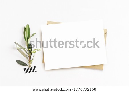 Feminine stationery, desktop mock-up scene. Blank horizontal greeting card, craft envelope and washi tape with green olive branch.White table background. Flat lay, top view. No people.