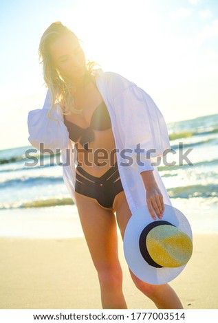 Carefree female tourist wearing bikini and shirt standing with straw sunhat on sandy beach against waving sea on sunny day in summer
