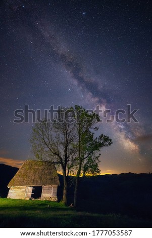 Old abandoned barn farm house with two trees next to it shot at night against a starry sky with milky way galactic core seen above shot in Dumesti, Salciua, Alba County in Romania